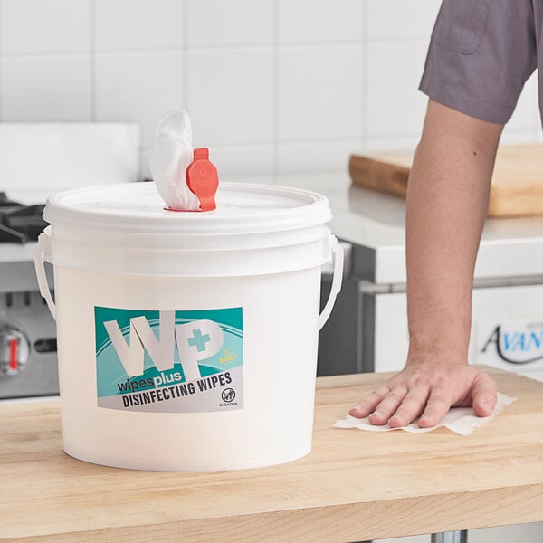 A person cleaning a surface with a white bucket of WipesPlus Lemon Scent alcohol free disinfecting wipes.