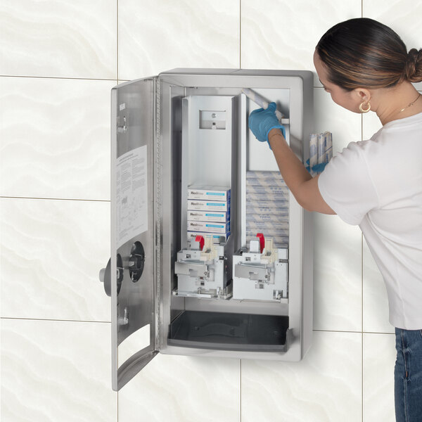 A stainless steel surface-mounted napkin and tampon dispenser with an open door, being refilled by a person wearing gloves.