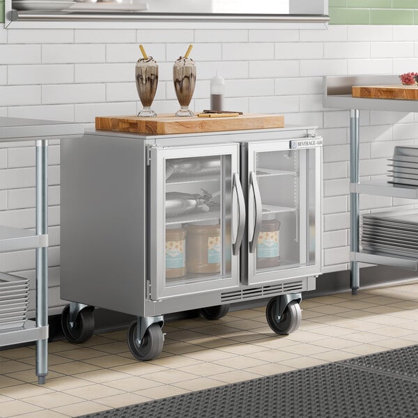A stainless steel undercounter refrigerator with glass doors and LED lighting on casters.