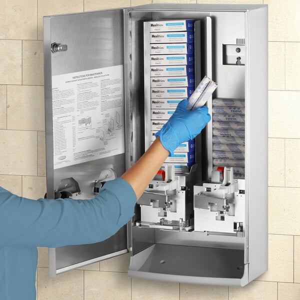 A woman in blue gloves is opening a metal cabinet holding a Bobrick sanitary napkin and tampon vendor.
