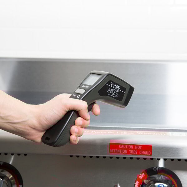 A hand holding a Taylor digital laser infrared thermometer over a stove.