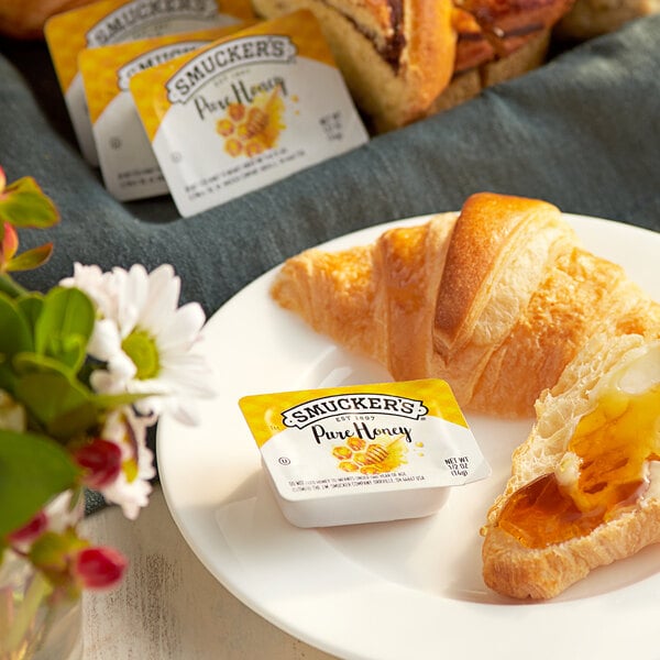 A plate with a croissant and a Smucker's honey portion cup on a table.