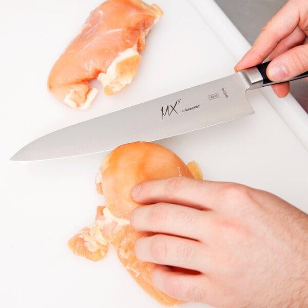 A person using a Mercer Culinary Japanese Gyuto knife to cut raw chicken on a cutting board.