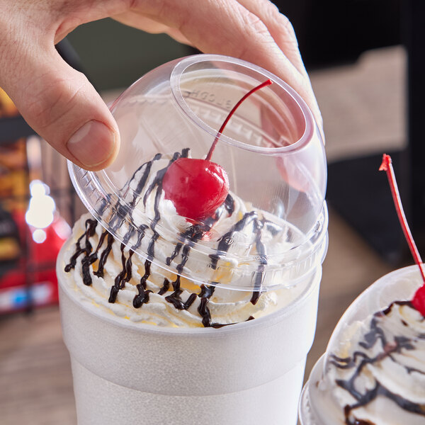 A clear plastic dome lid with a 2-inch hole being placed on a cup topped with whipped cream, chocolate drizzle, and a cherry.