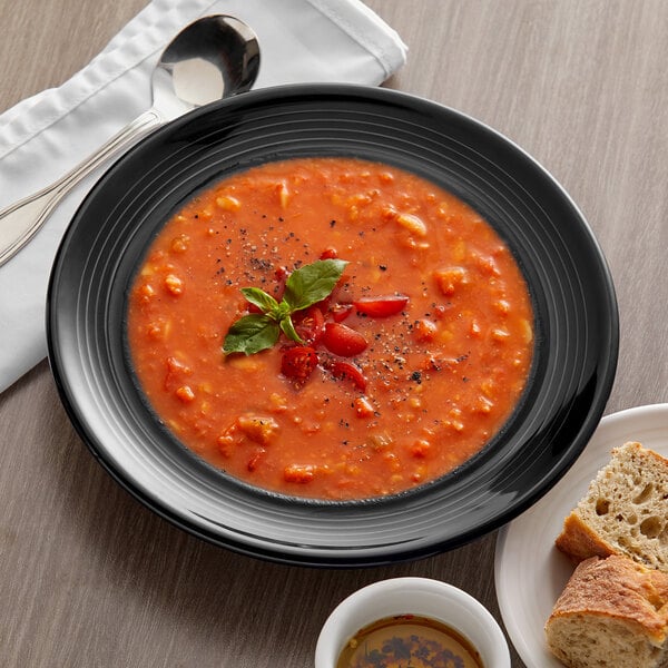 A Tuxton black china bowl filled with soup with a spoon and bread on a plate.