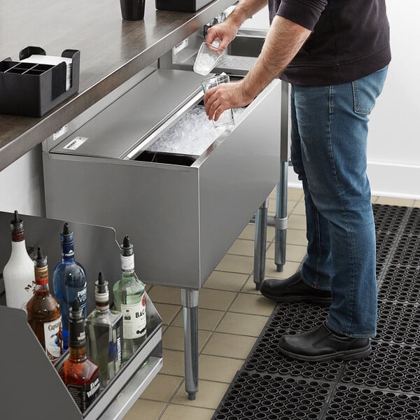 A person is scooping ice from a stainless steel ice bin with a sliding lid, located under a countertop in a bar setting.