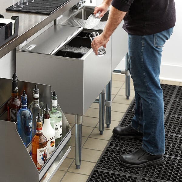 A person is scooping ice from a stainless steel ice bin with a sliding lid in a bar setting, with bottles of liquor visible nearby.