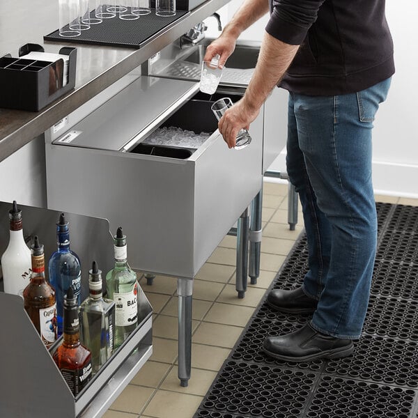 A person is scooping ice from a stainless steel ice bin with a sliding lid in a bar setting.
