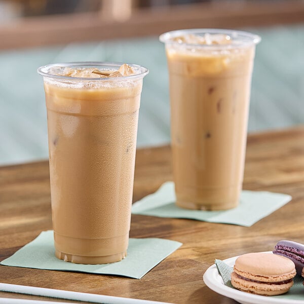 Two Choice clear plastic cups filled with iced coffee on a wooden table with cookies.
