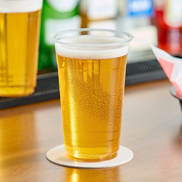 A clear plastic cup of beer with foam on a table.