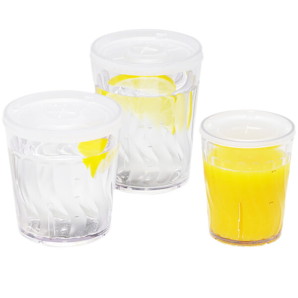 Three Dinex clear plastic tumblers filled with orange juice and garnished with lemon slices on a table with a bowl of lemons.
