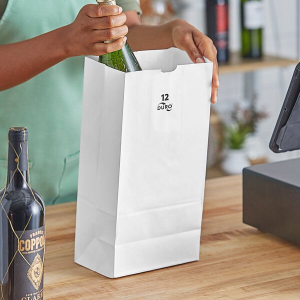 A person opening a bottle of wine and pouring it into a white Duro paper bag.