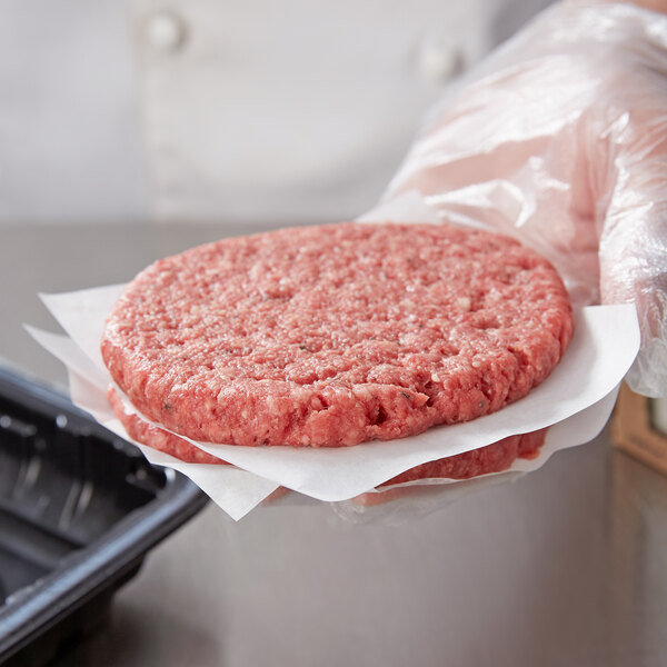 A hand holding a raw hamburger with a black container of Choice patty papers on a counter.