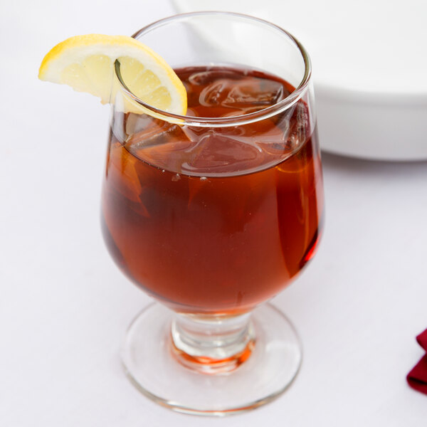 A Libbey footed goblet filled with brown liquid and a lemon slice on the rim.