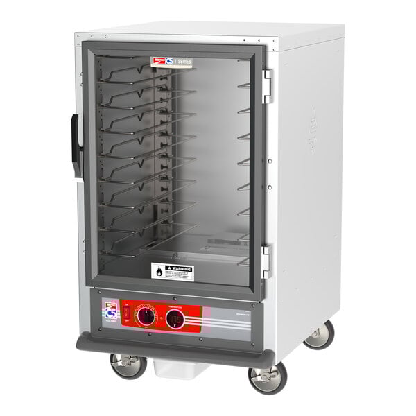 A large white holding cabinet with a clear glass door on wheels.