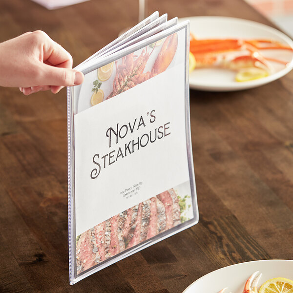 A hand holding a H. Risch, Inc. clear menu cover with a plate of food on a table.