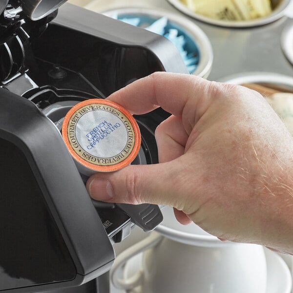 A hand holding a round orange and black Caffe de Aroma French Vanilla Cappuccino single serve cup.