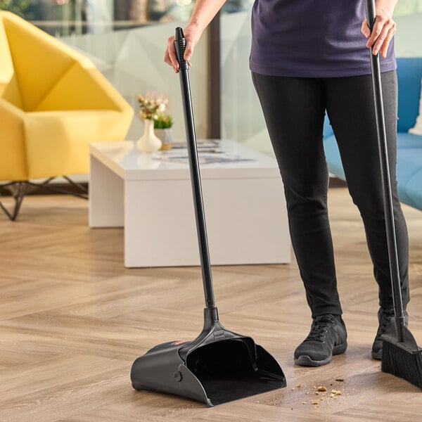 A black plastic upright dust pan with a long handle being used to collect debris from a floor.