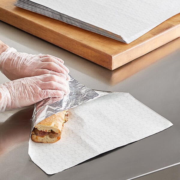 A pair of gloved hands wrapping a sandwich with Choice Insulated Foil Sheets.