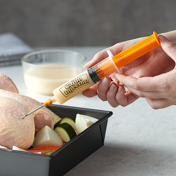 A hand using a Cajun Injector syringe to inject yellow marinade into a piece of chicken.