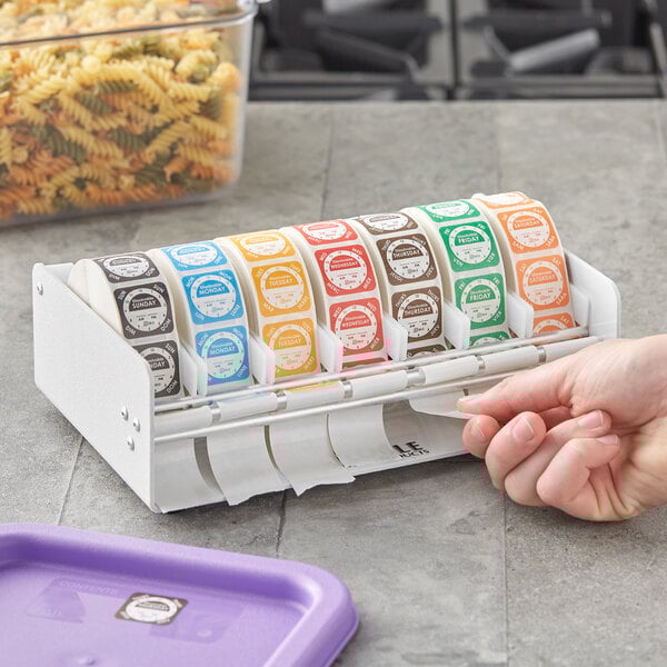 A hand using a Noble Products white plastic label dispenser to hold a roll of food labels.