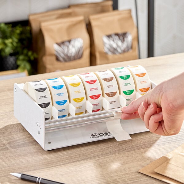 A white plastic label dispenser with seven slots, each holding a roll of day-of-the-week labels.