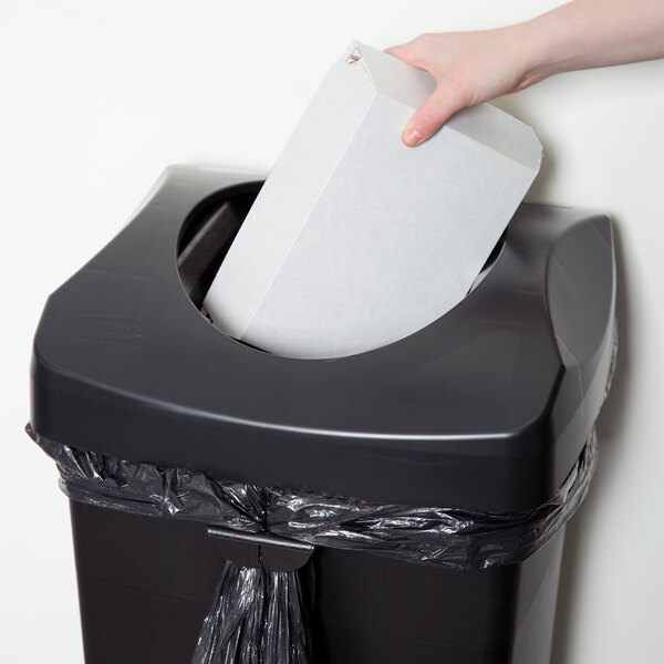 A hand putting a white box into a Rubbermaid black square commercial trash can.