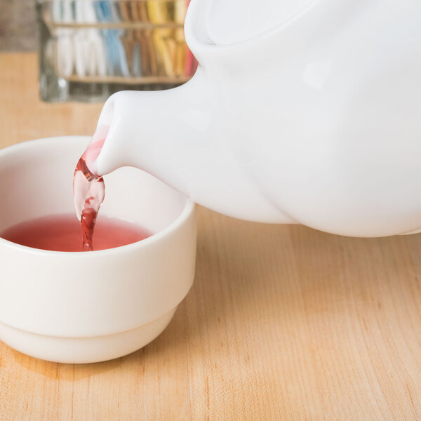 A white Tuxton china teapot pouring red liquid into a white cup.