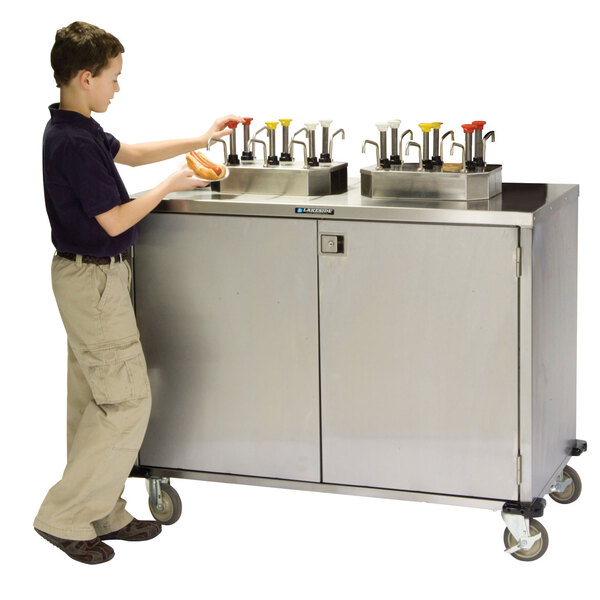 A boy standing next to a Lakeside stainless steel condiment cart.