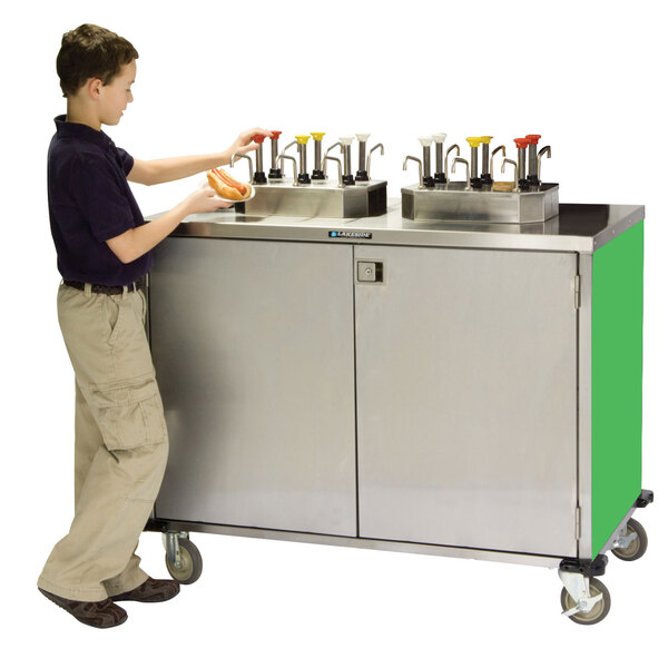 A young boy standing next to a Lakeside stainless steel condiment cart with green accents.