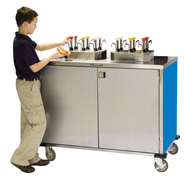 A young boy standing next to a Lakeside stainless steel condiment cart.