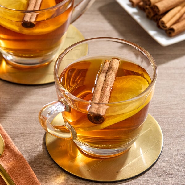 A glass cup of tea garnished with a cinnamon stick and a slice of orange, placed on a gold coaster.