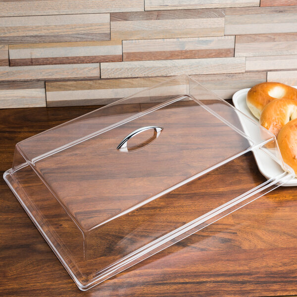 A clear Cal-Mil rectangular tray cover on a wood surface with a plate of bagels inside.