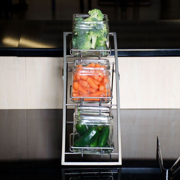An American Metalcraft chrome three-tier display stand holding glass jars of vegetables.