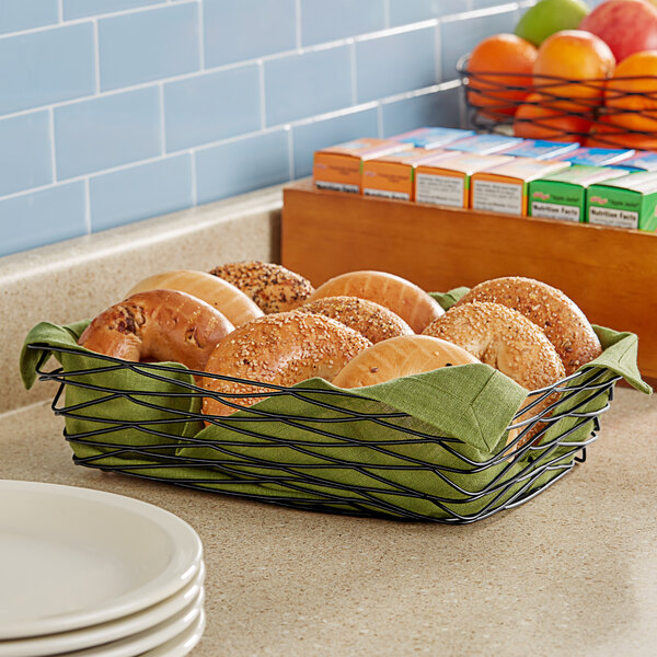 A Tablecraft rectangular black wire basket holding a bagel and fruit on a counter.