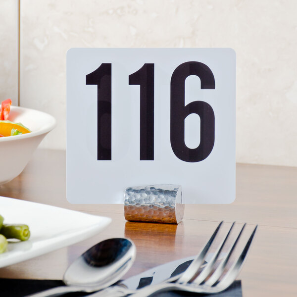 An American Metalcraft hammered aluminum cylinder table card holder with a white card and black numbers on a table.