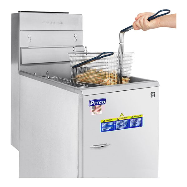 A hand holding a basket of fries in front of a Pitco stainless steel floor fryer.