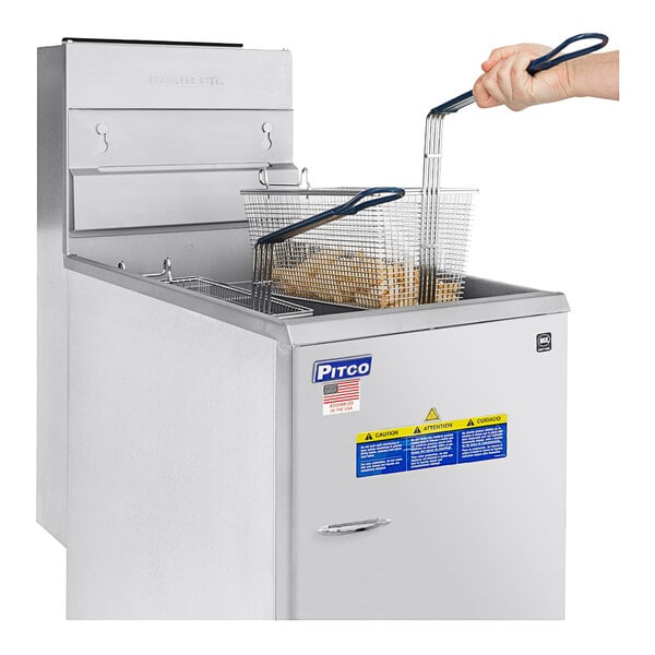 A hand using tongs to hold a basket of fries in front of a Pitco stainless steel floor fryer.