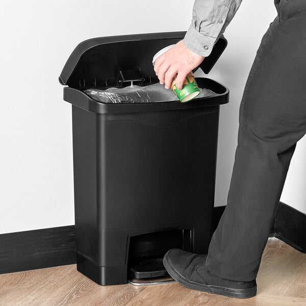 A hand putting a green and yellow cup into a black Rubbermaid trash can.