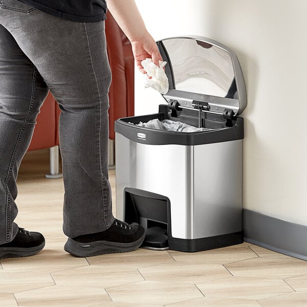 A woman in jeans using a Rubbermaid Slim Jim trash can to put a plastic bag into a trash can.