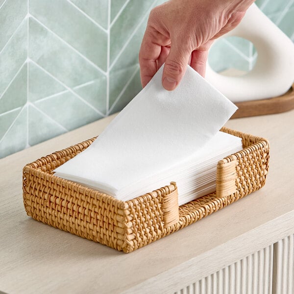 A stack of white linen-feel guest towels in a woven basket with a hand pulling one towel from the pack.