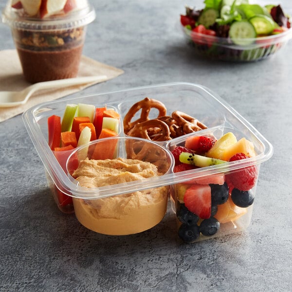 An assortment of pretzels, fruit, vegetables, and hummus in a PLA plastic container