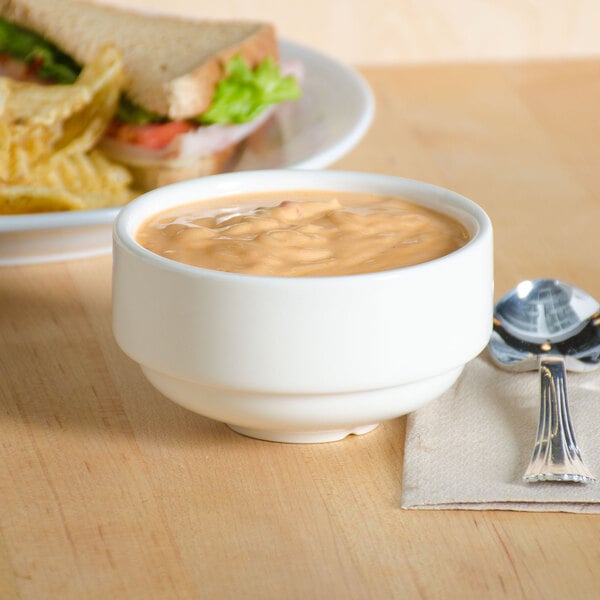 A Tuxton eggshell china soup cup filled with soup next to a plate of food on a table.