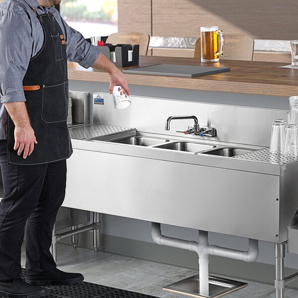 A man in a black apron standing in front of an Advance Tabco 3 compartment underbar sink with drainboards.