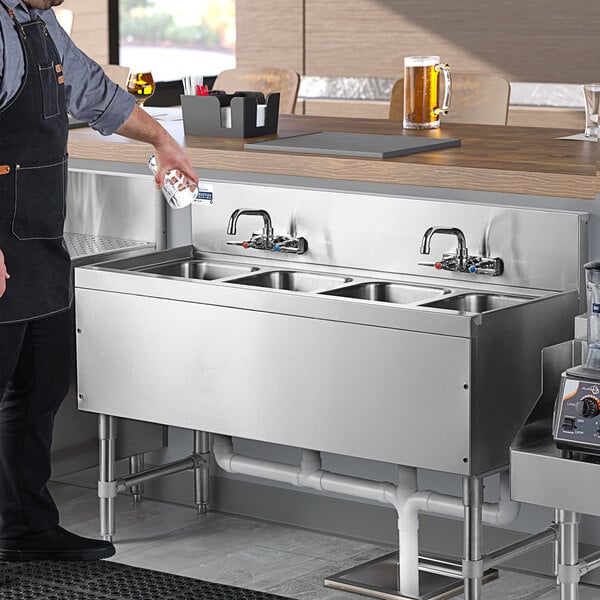 A man in a black apron pouring water into an Advance Tabco stainless steel underbar sink with faucets.