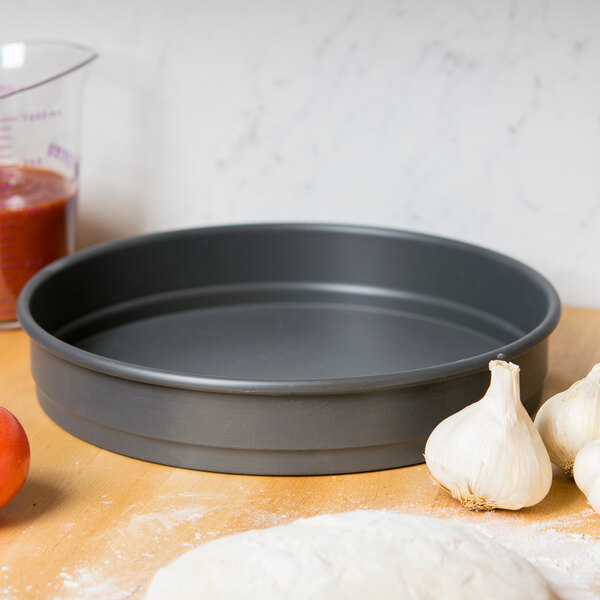 An American Metalcraft Hard Coat Anodized Aluminum pizza pan with pizza dough and ingredients on a table.
