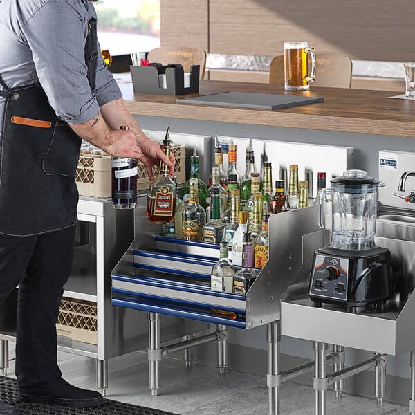 A man in an apron placing bottles of alcohol on an Advance Tabco Prestige Series stainless steel liquor display rack.
