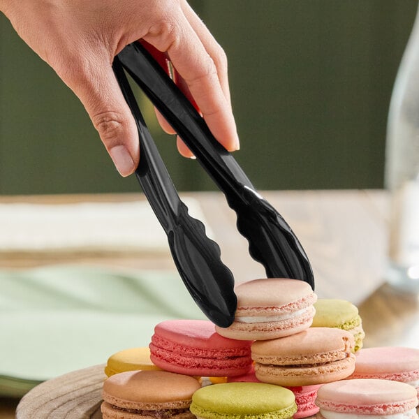 A pair of black disposable polypropylene tongs being used to pick up a macaron from a plate.