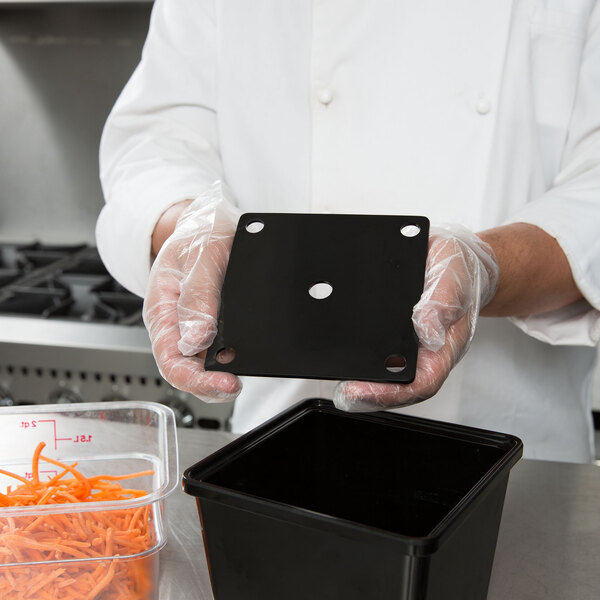 A hand holding a black square GET melamine container over a bowl of carrots.