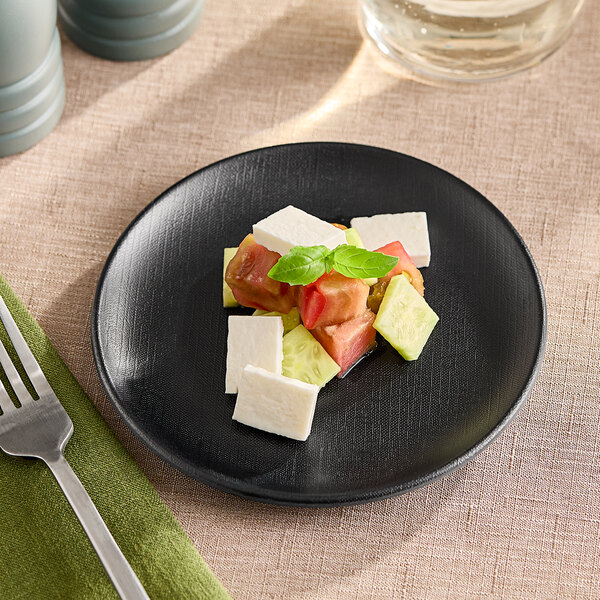 A Elite Global Solutions black melamine plate with a slice of tomato and a slice of cheese on a table with a green cloth.